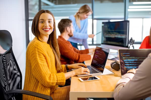 Smiling woman in a yellow sweater working on a laptop, surrounded by colleagues collaborating in a modern office setting, illustrating teamwork and digital marketing strategies.