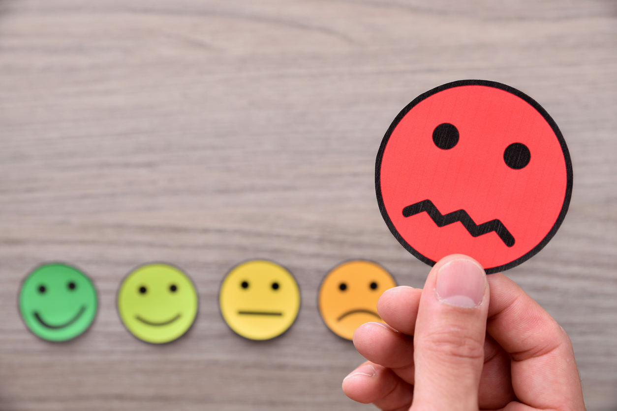 Hand holding a red unhappy face icon, with a gradient of five emoticons ranging from green happy to red unhappy, symbolizing client dissatisfaction in digital marketing.
