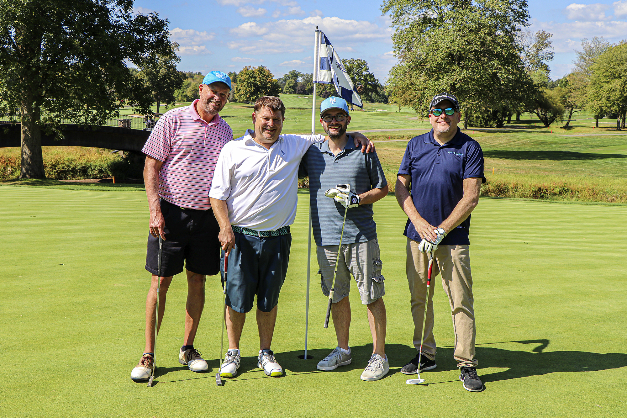 Four men posing on a golf course during a charity event, smiling and holding golf clubs, with a flag in the background and trees lining the fairway.