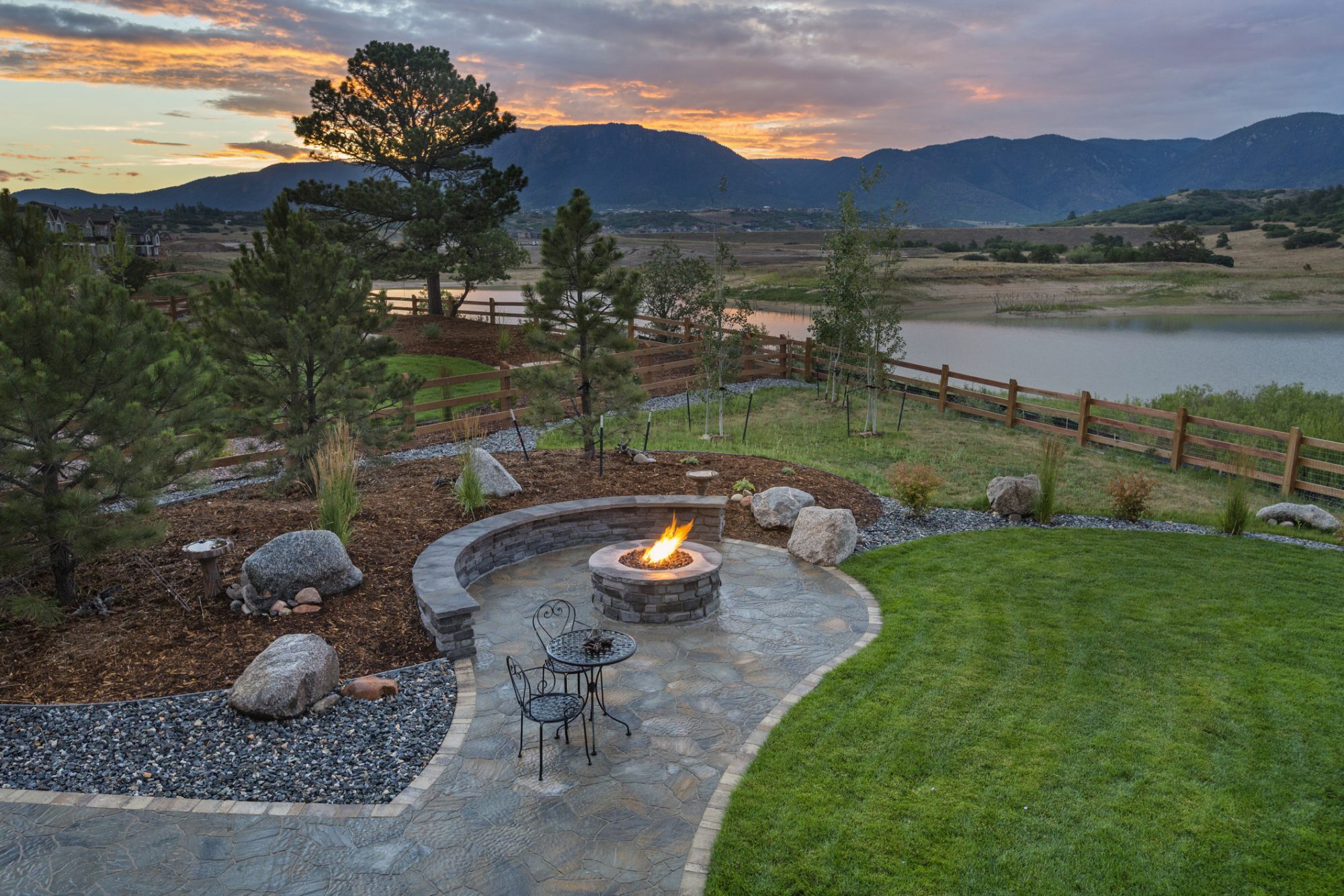Scenic landscape featuring a stone fire pit surrounded by a circular patio, chairs, and lush greenery, with mountains and a lake in the background, showcasing ideal outdoor living spaces for landscaping marketing.